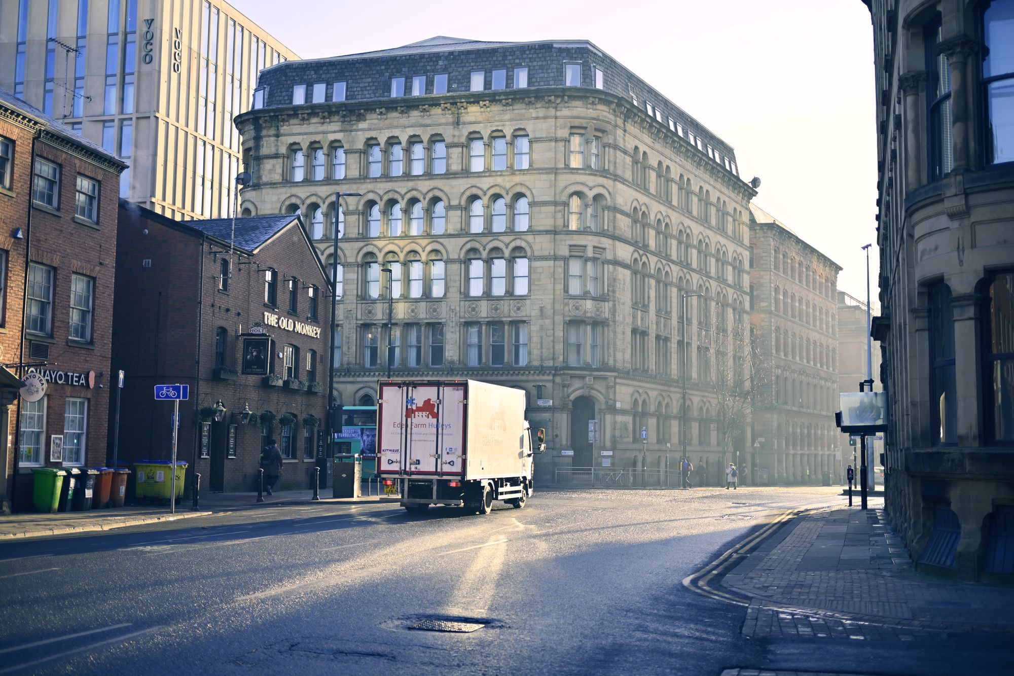 A quiet Manchester street in early morning light, wet road surface reflecting the stone buildings along it.