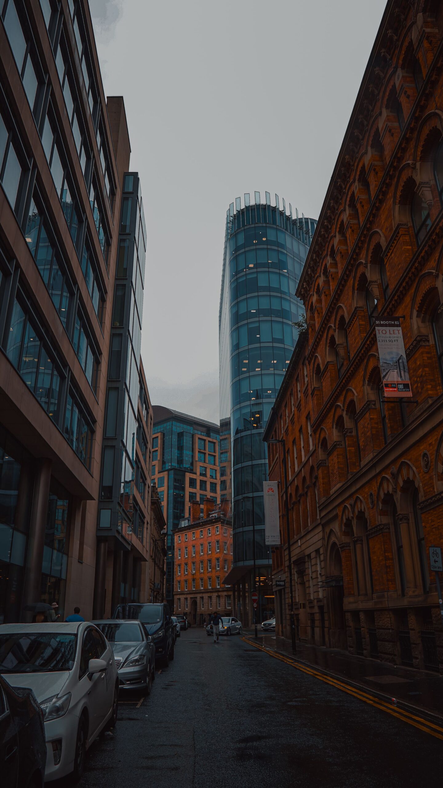 Looking up a rain-soaked Manchester street where red brick Victorian frontages meet contemporary glass towers.