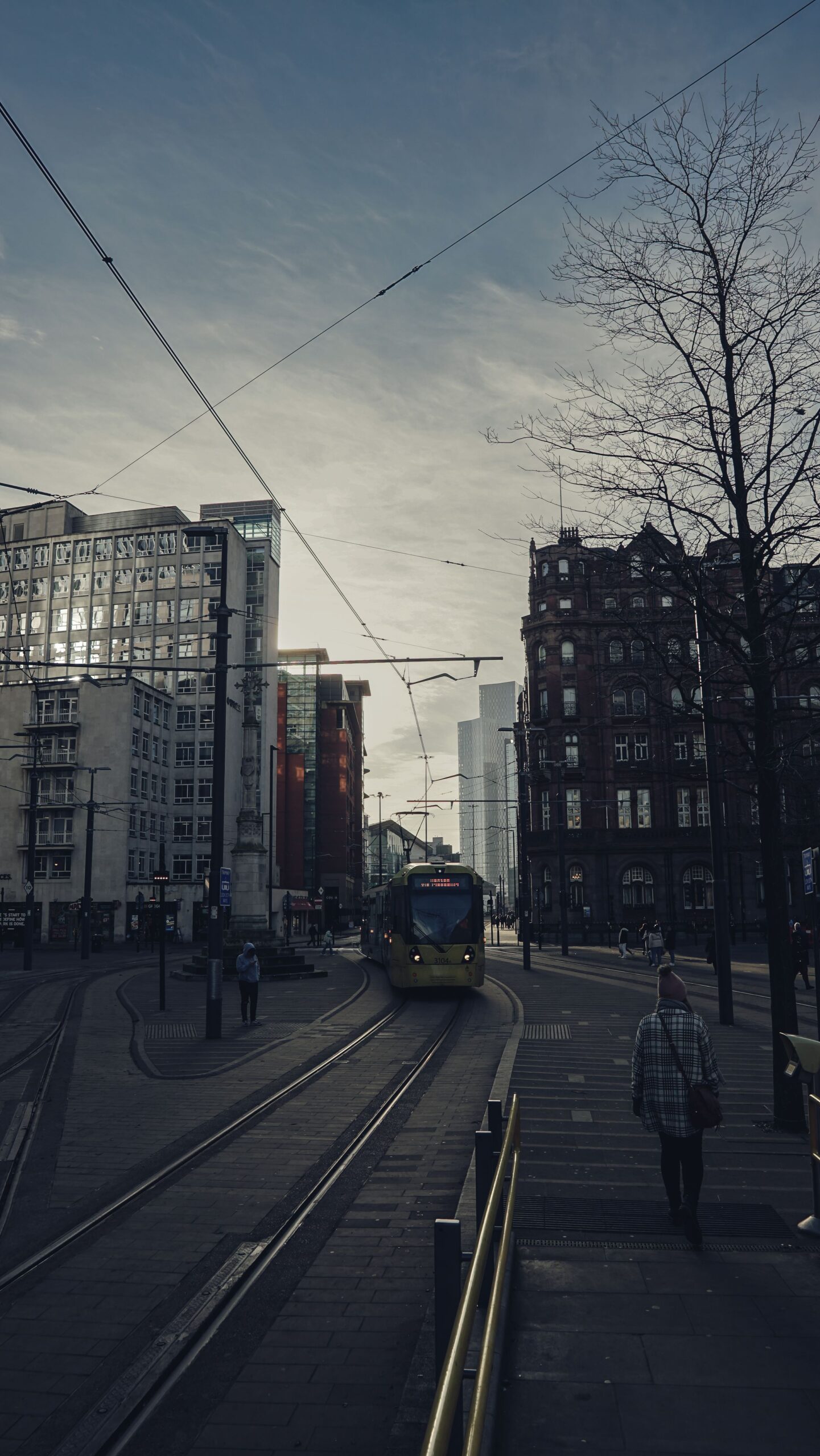 A Metrolink tram moving through central Manchester at twilight, with Victorian and modern buildings either side of the track.